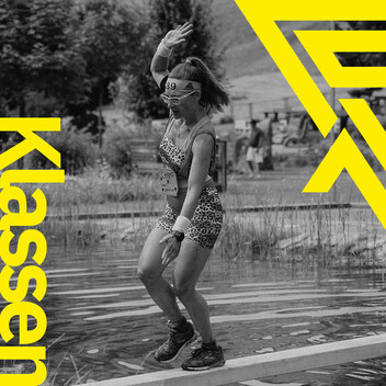 Zu den Klassen und Wertungen Frau in schriller Sportkleidung balanciert auf einem schmalen Balken über einem Teich. Arlberger Wadlbeisser Logo als Designelement in gelb am rechten Bildrand und Schriftzug "Klassen" vertikal am linken Bildrand. Bild in Graustufen.